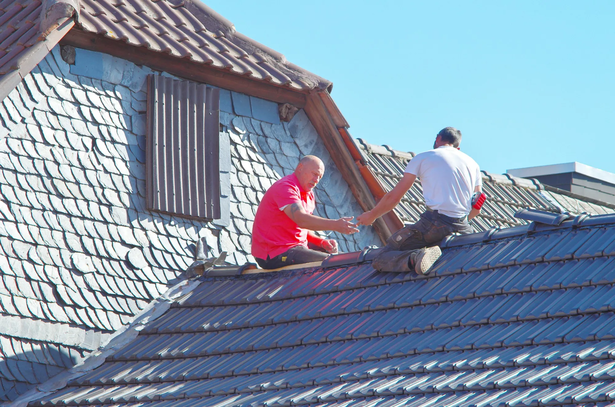 Two roofers repairing a steep slate roof on a sunny day